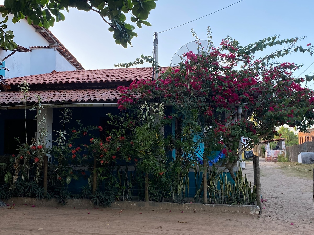 A colorful exterior view of a house is framed by vibrant flowering plants, showcasing a rustic tiled roof. The walls are painted in soothing tones, and a dirt pathway leads towards the entrance. Surrounding greenery adds to the natural charm of the setting.