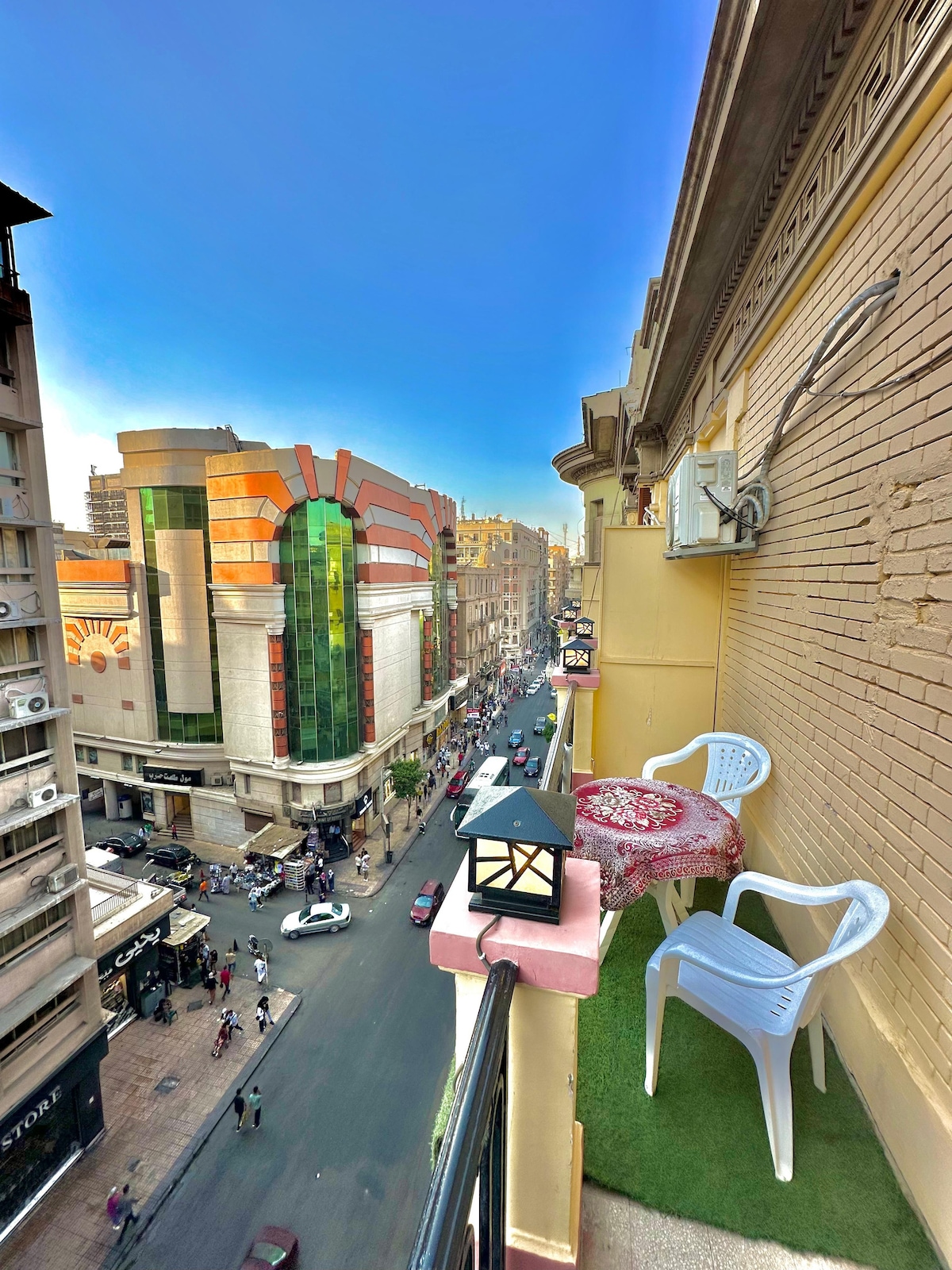 A balcony offers a view of a bustling street, characterized by colorful storefronts and a clear sky. Two white chairs are positioned on a green patch of artificial grass, complemented by a small table adorned with a decorative cloth. A lantern adds to the setting.
