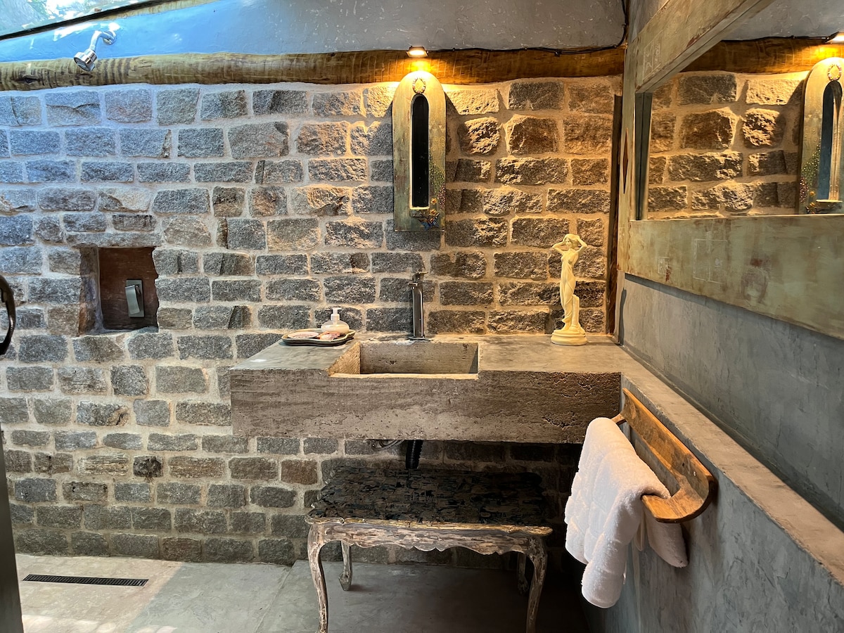 A unique bathroom features exposed stone walls and a sleek concrete sink. A small statue decorates the counter, while a towel is neatly hung on a wooden rack. Soft lighting is provided by wall-mounted fixtures, enhancing the space's calming ambiance.