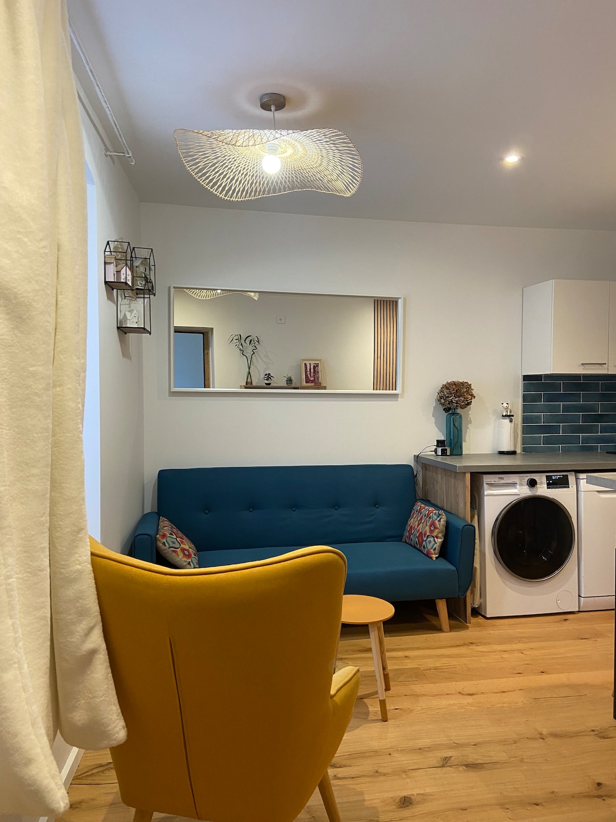A cozy living area features a blue sofa adorned with decorative cushions, paired with a vibrant yellow armchair. A small round table sits in front of the sofa, and a large mirror reflects the soft lighting from the stylish ceiling fixture. A washing machine is discreetly positioned nearby.