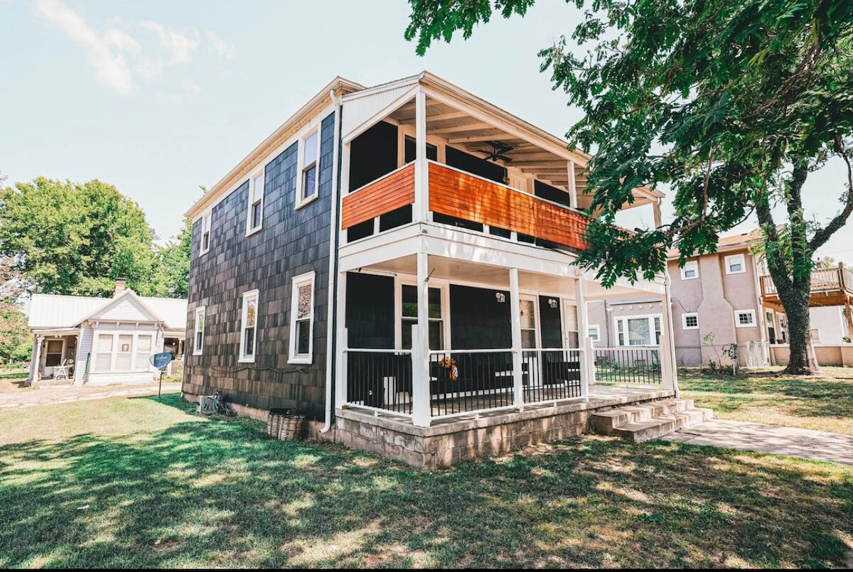 A two-story apartment is set within a landscaped area, featuring a modern exterior with dark shingles and a light wood balcony. The spacious porch is supported by white railings, providing an inviting outdoor space. Lush green grass surrounds the property.