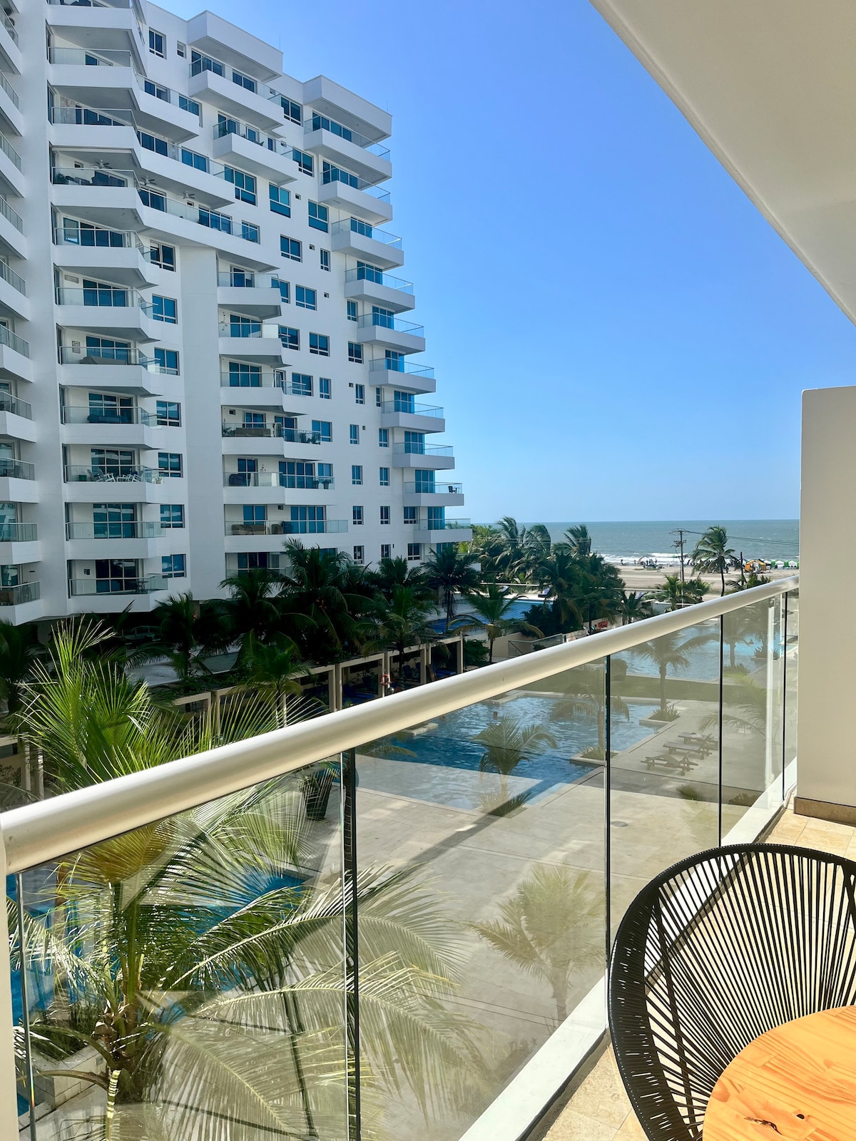 A bright balcony view showcases palm trees framing the scene, with a glimpse of the ocean in the distance. Below, a large swimming pool is visible against the backdrop of a contemporary building structure, all under a clear blue sky.