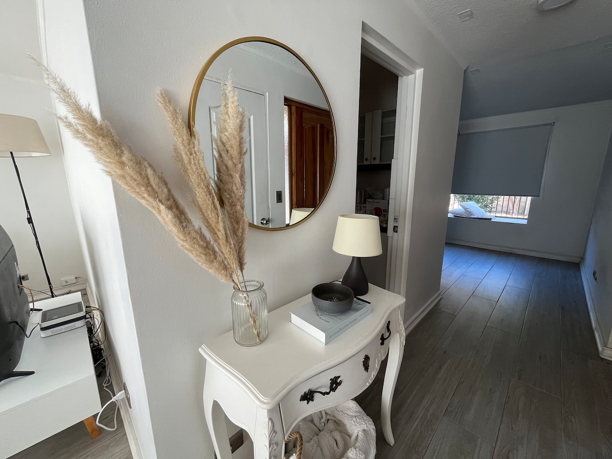A hallway is presented, showcasing light-colored walls and a wooden floor. A round mirror hangs above a console table, which displays a glass vase with decorative grasses and a small lamp. A doorway leads to a room with a window, providing a glimpse of natural light.