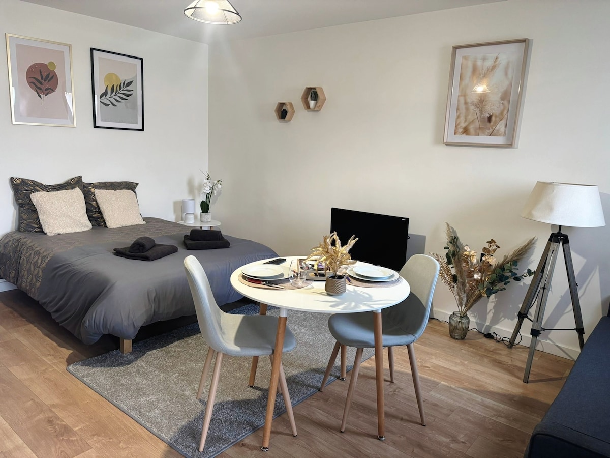 A spacious studio features a cozy sleeping area with a double bed adorned with neutral linens. A dining table for two is set against the backdrop of decorative wall art. Soft lighting is provided by a standing lamp, enhancing the welcoming ambiance.