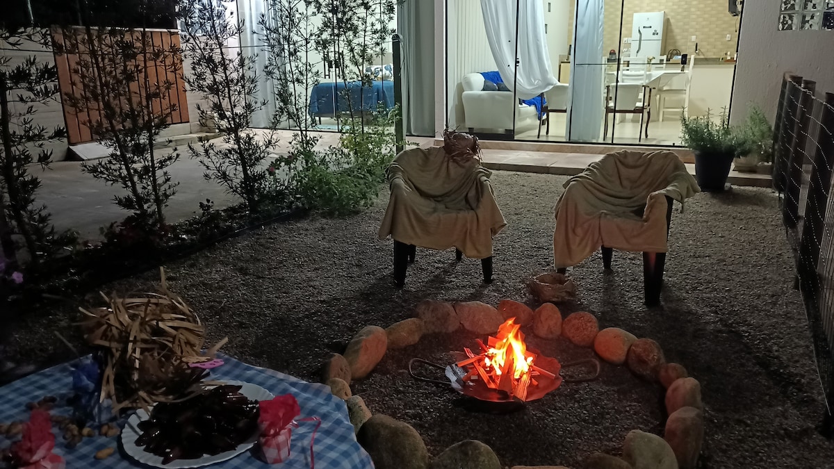 An outdoor area features a circular stone fire pit surrounded by comfortable chairs, draped with soft coverings. A checkered tablecloth is placed nearby with items for gatherings. The backdrop includes softly illuminated doors leading into the home's interior, enhancing a cozy atmosphere.
