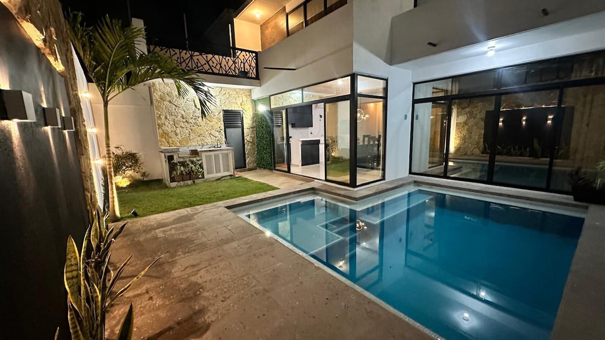 A private pool is framed by natural stone and glass walls, creating an inviting outdoor space. Lush green plants sit along the edges, while the soft lighting enhances the tranquil evening ambiance. A well-maintained grassy area complements the pool, offering a relaxing retreat.