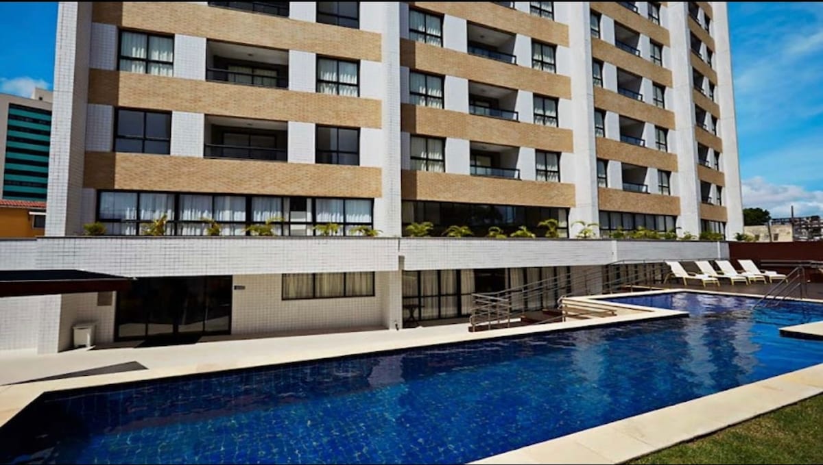 An outdoor swimming pool showcases a sleek design, featuring blue tiles that reflect the sky above. Surrounding lounge chairs provide space for relaxation. The building's modern exterior, comprised of tan and white panels, adds a contemporary touch to the overall setting.