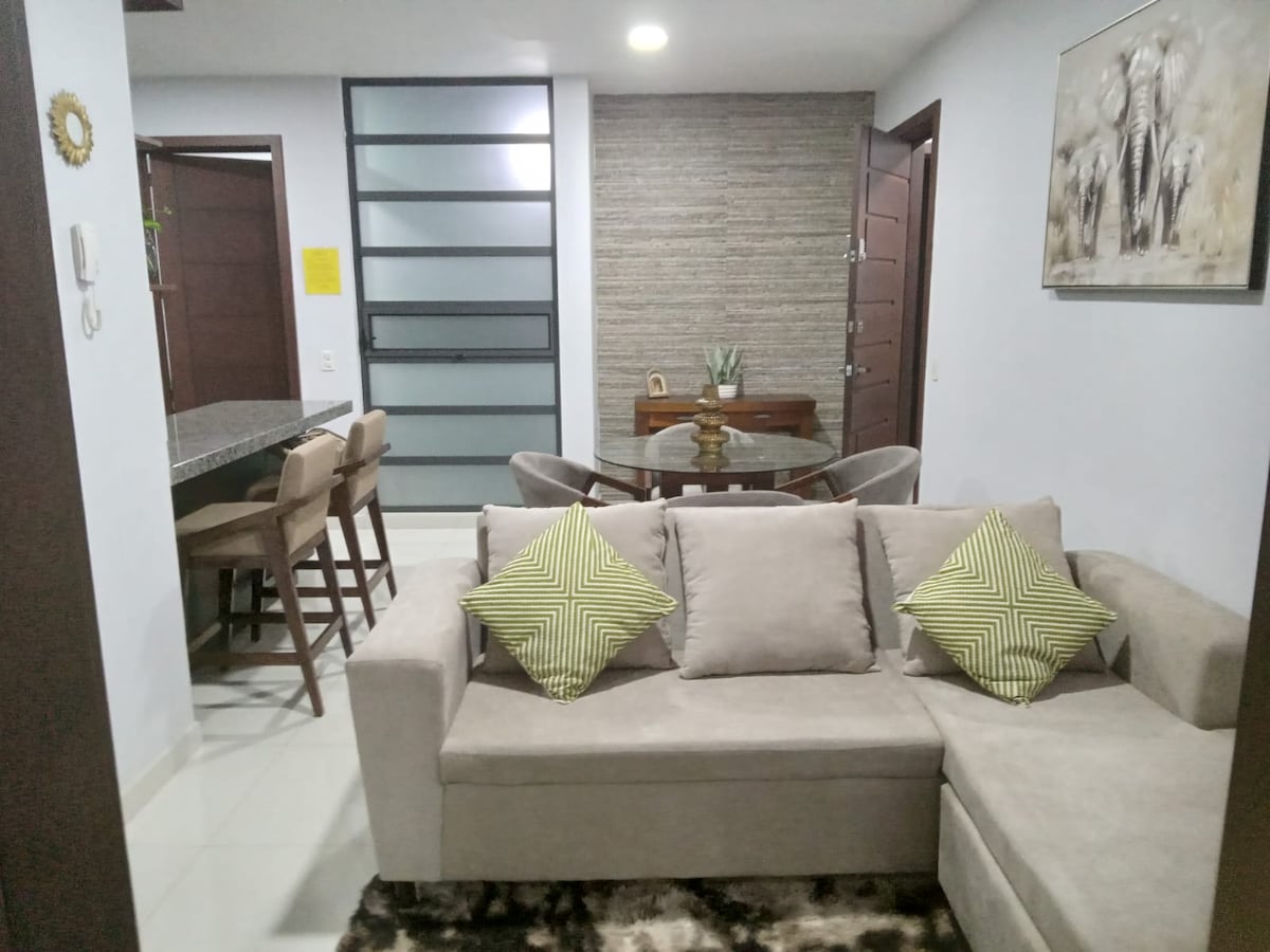 A contemporary living space features a light-colored sectional sofa adorned with patterned cushions. A dining table with four chairs is visible in the background, complemented by a kitchen bar with additional seating. Neutral tones and natural light from nearby windows create a welcoming ambiance.