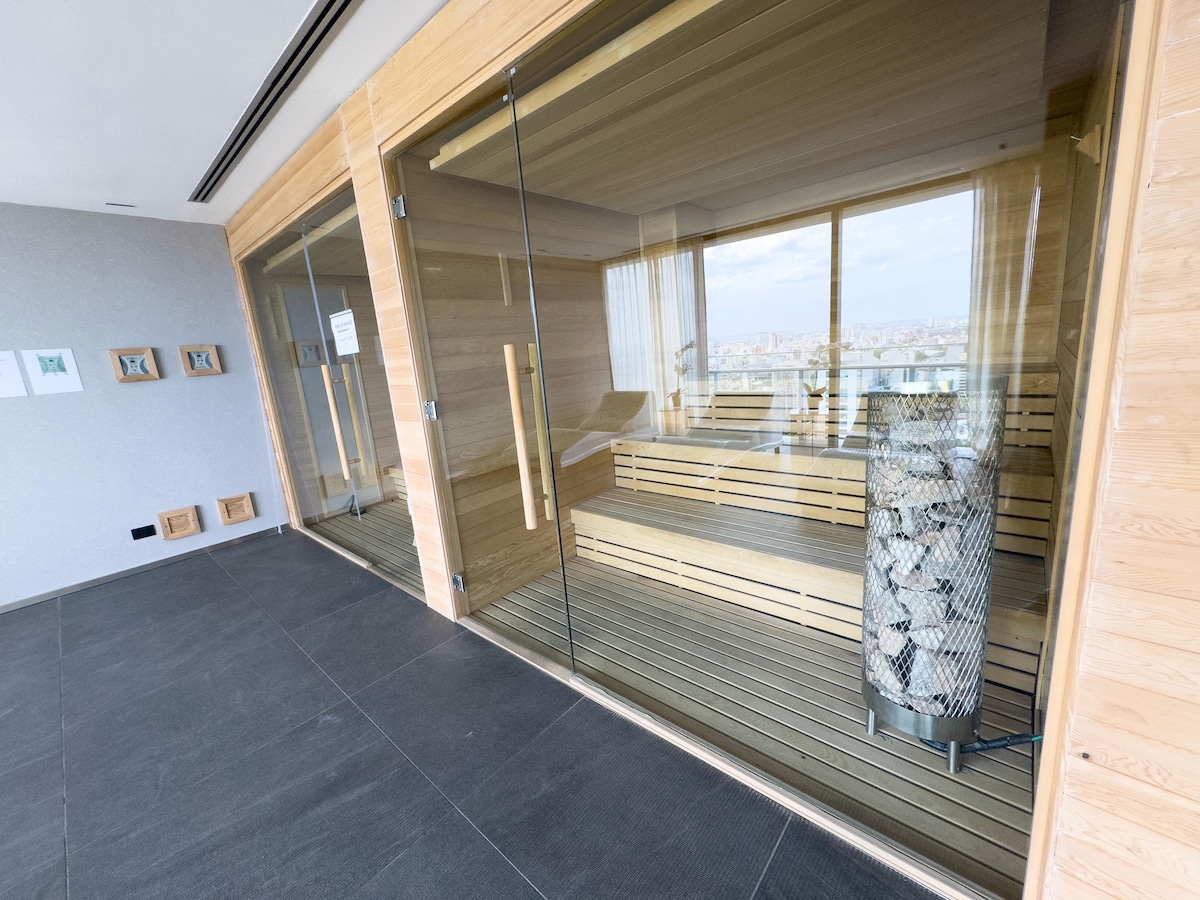 A modern sauna room is enclosed with transparent glass walls, allowing views of the cityscape. Inside, wooden benches provide seating, complemented by a decorative stone feature. Large windows invite natural light, contributing to a bright and welcoming atmosphere.