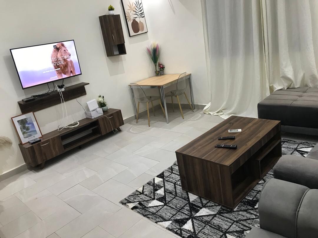 A modern living area is presented with a flat-screen television mounted on a wooden console. A rectangular coffee table is centrally placed on a patterned rug, surrounded by a sectional sofa. A dining table with transparent chairs is located against the wall, illuminated by natural light from the windows.