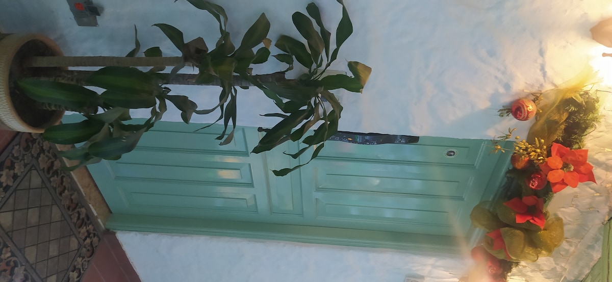 A green door is framed by potted plants and decorative elements, including seasonal decor. A tiled area with intricate patterns is visible, enhancing the welcoming entrance. Soft lighting casts a warm glow on the surrounding features.