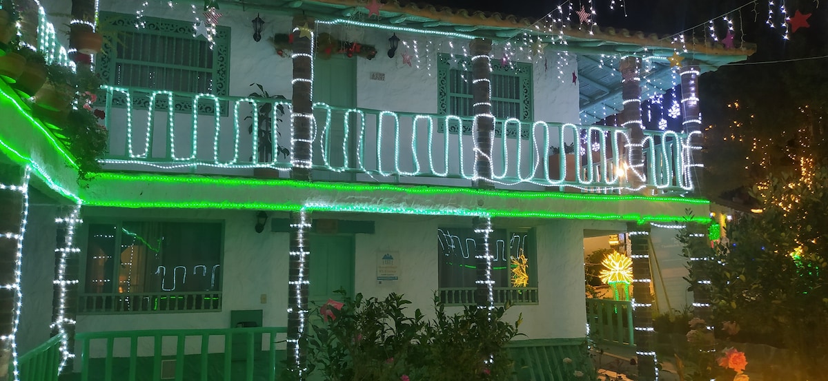 A charming multi-level building is adorned with colorful holiday lights, illuminating the green railings and exterior. Potted plants are placed throughout, enhancing the lively atmosphere. A small garden area is visible in the foreground, contributing to the welcoming ambience of the property.
