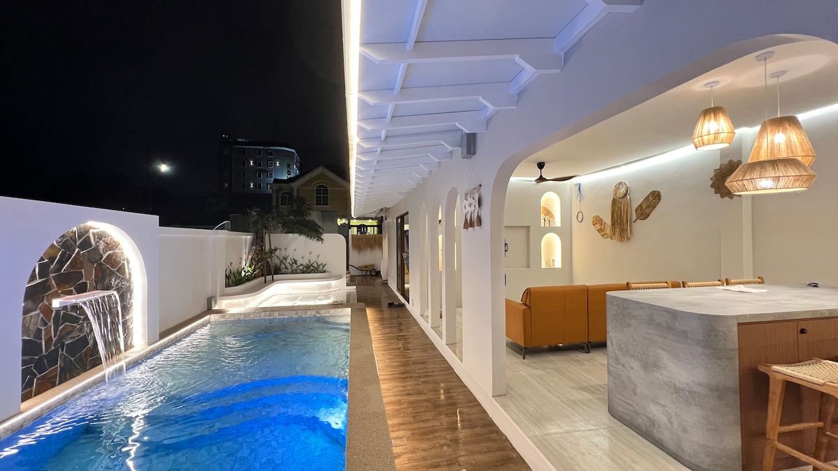 An illuminated pool area is seen at night, featuring a serene swimming pool with a gentle waterfall effect. The adjacent open space includes a stylish kitchen island and a seating area adorned with woven pendant lights, creating a warm ambiance under the soft lighting.