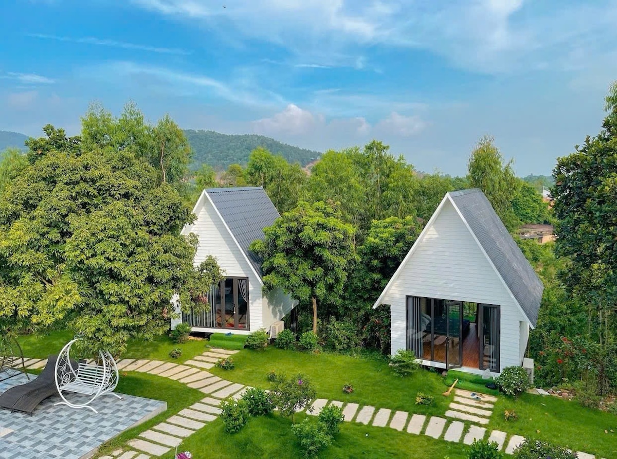 Two modern cottages with white exteriors are situated among lush greenery, framed by rolling hills in the background. A stone pathway leads to each cottage, while a hanging chair is visible on the side. The scene reflects a serene outdoor environment.