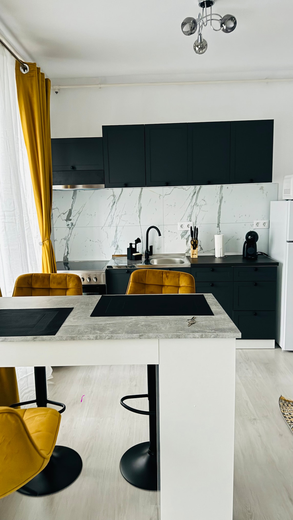 A modern kitchen space features sleek dark cabinetry paired with a light marble backsplash. Yellow accent chairs surround a concrete table, creating a stylish dining area. Natural light filters through sheer white curtains, enhancing the simplicity and functionality of the layout.