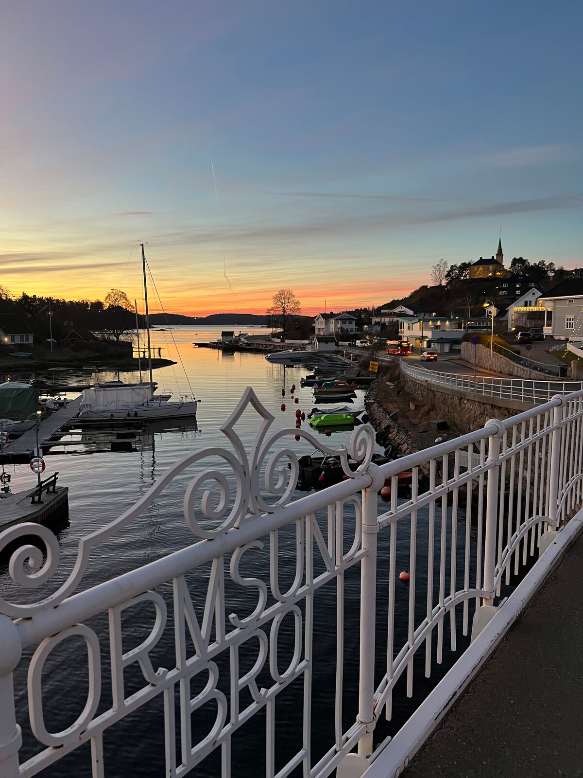 A view of the harbor at sunset features calm water reflecting vibrant hues. Boats are moored along the shore, with buildings and trees silhouetted against the skyline. A decorative white railing with text lines the sidewalk, adding a charming touch to the coastal scene.
