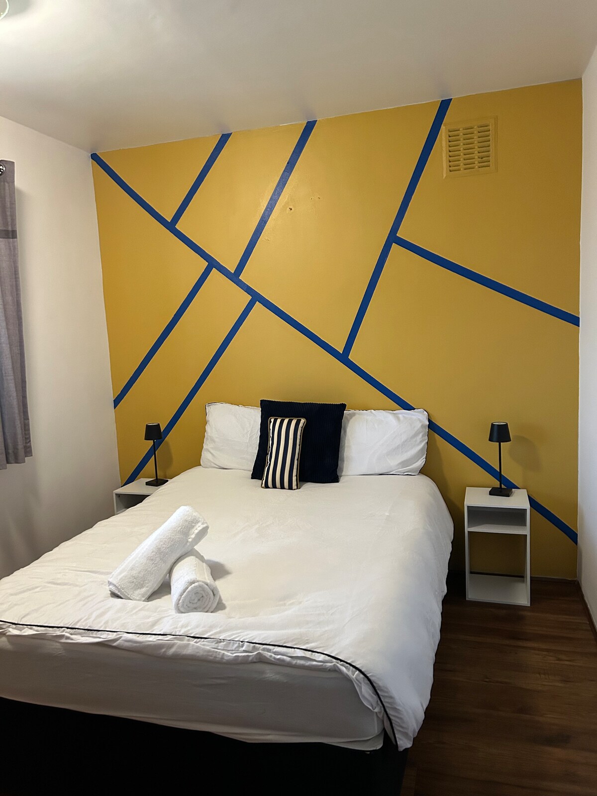 A bedroom features a spacious bed with white bedding, complemented by two nightstands, each holding a small lamp. The yellow accent wall is adorned with intersecting blue lines, adding a modern touch. Fresh towels are neatly arranged at the foot of the bed.