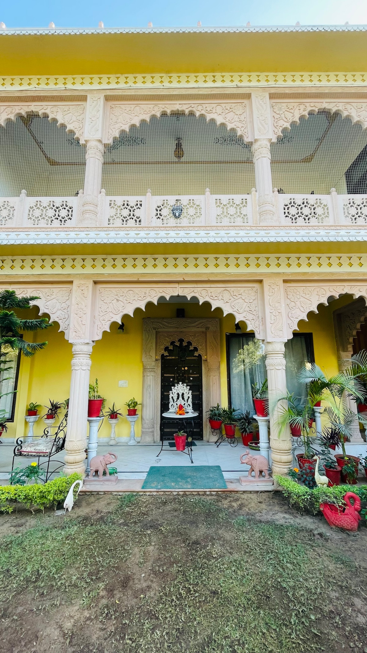 The entrance features ornate yellow façades complemented by intricate architectural details. Two decorative elephants are positioned near the pathway, which leads to an inviting entrance adorned with potted plants. A central idol is visible, framed by detailed arches and a welcoming atmosphere.