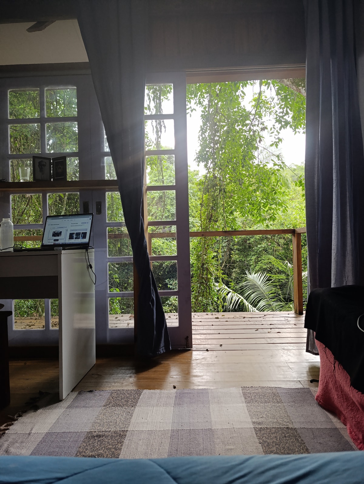 A serene view from a suite showcases large glass doors opening to a private balcony, surrounded by lush greenery. Natural light filters in, highlighting a small workspace with a laptop. A soft rug covers the wooden floor, enhancing the cozy ambiance.