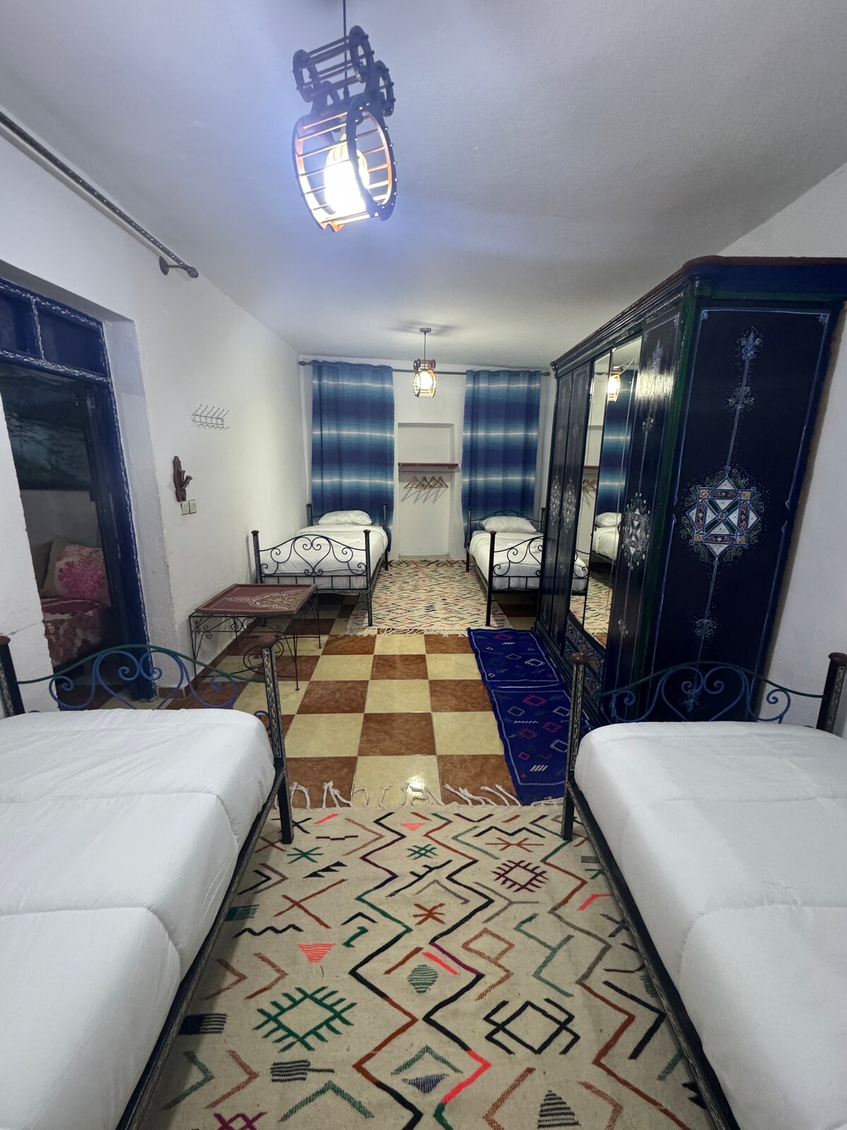 A vibrant room featuring four iron beds arranged for comfort. The floor showcases a colorful geometric pattern, complemented by a decorative area rug. Two large windows are framed with blue curtains, and a lantern-style light fixture is suspended from the ceiling.