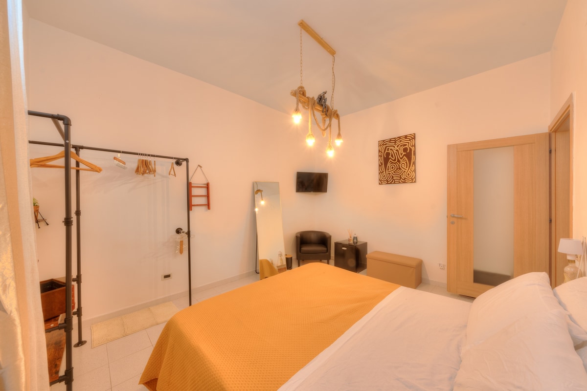 A well-lit bedroom features a spacious bed covered with a yellow blanket. A mirror stands adjacent to the bed, and a TV is mounted on the wall opposite. Additional furnishings include a small seating area with two chairs and a storage bench, complemented by a clothing rack.