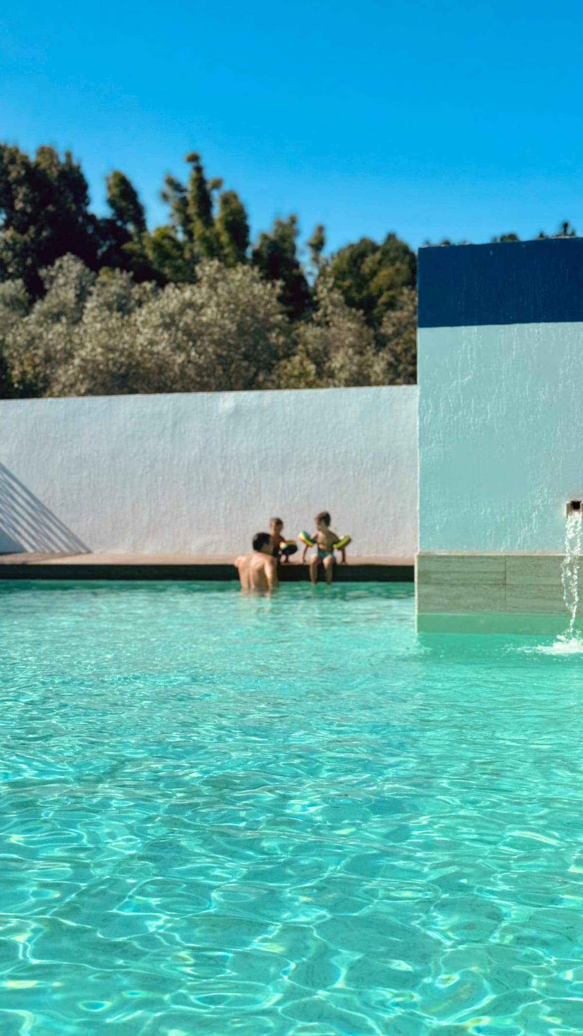 A crystal-clear swimming pool reflects sunlight, surrounded by a smooth, white wall adorned with greenery. Two individuals enjoy the water, providing a glimpse of relaxation and leisure in a tranquil outdoor setting.