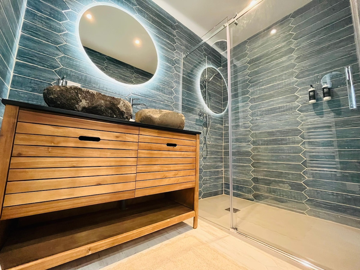 A modern bathroom features blue-toned wall tiles, creating a calming atmosphere. Two round mirrors are illuminated above a wooden vanity with natural stone basins. A glass shower enclosure is visible, showcasing a sleek design with minimalist fixtures.
