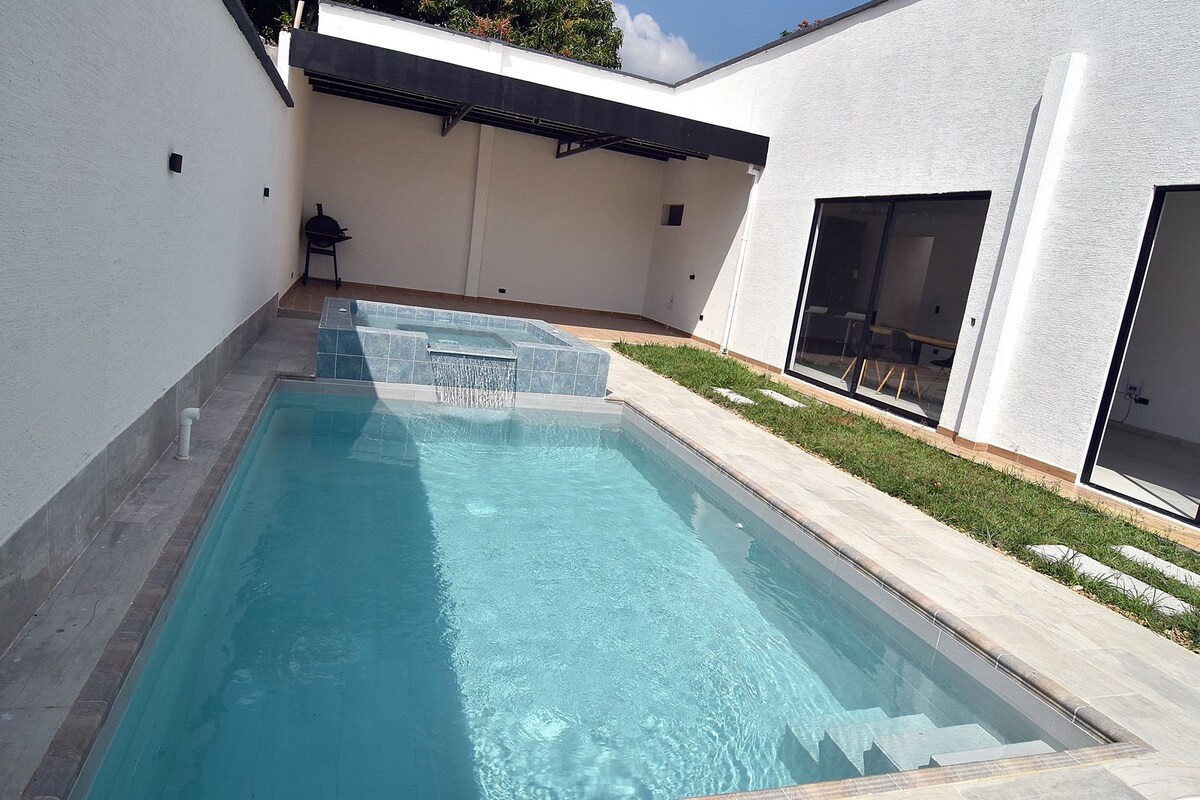 A private outdoor area features a clear pool, accompanied by a hot tub. The surroundings include a well-manicured lawn and an outdoor dining space, all enclosed by white walls, providing a serene environment for relaxation.
