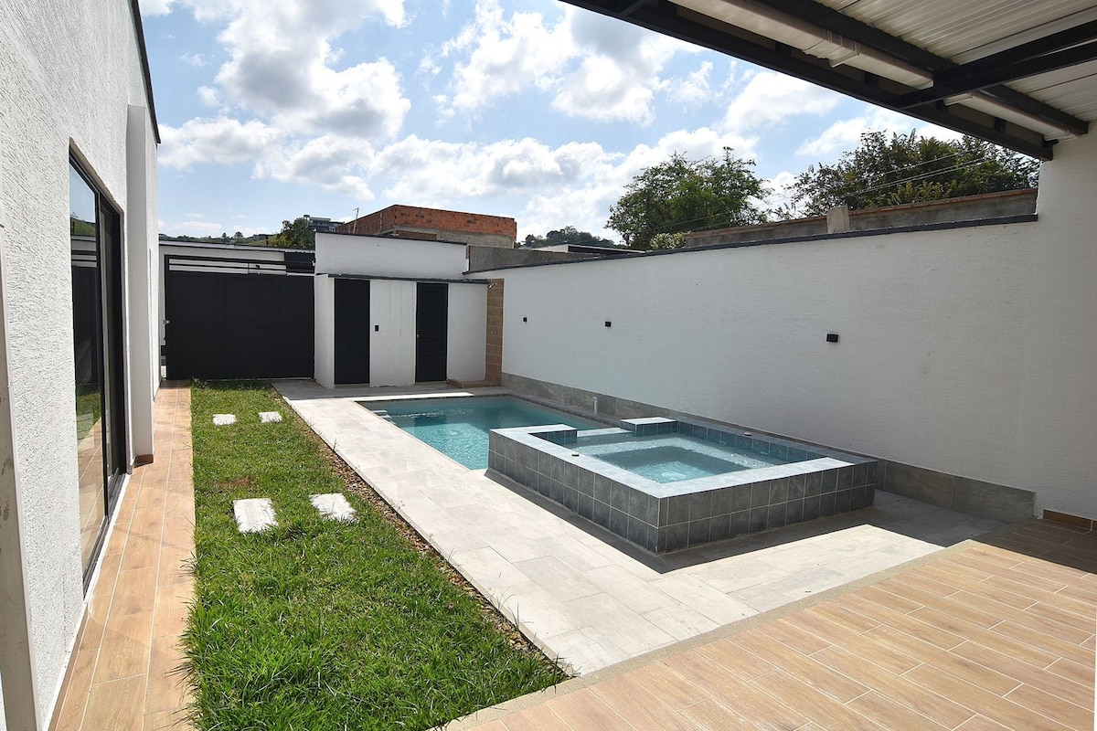 An outdoor space features a tranquil pool area surrounded by neatly cut grass. A smaller hot tub is positioned adjacent to a larger swimming pool. The area is enclosed by clean white walls, with a shaded terrace providing a comfortable space for relaxation and enjoyment.