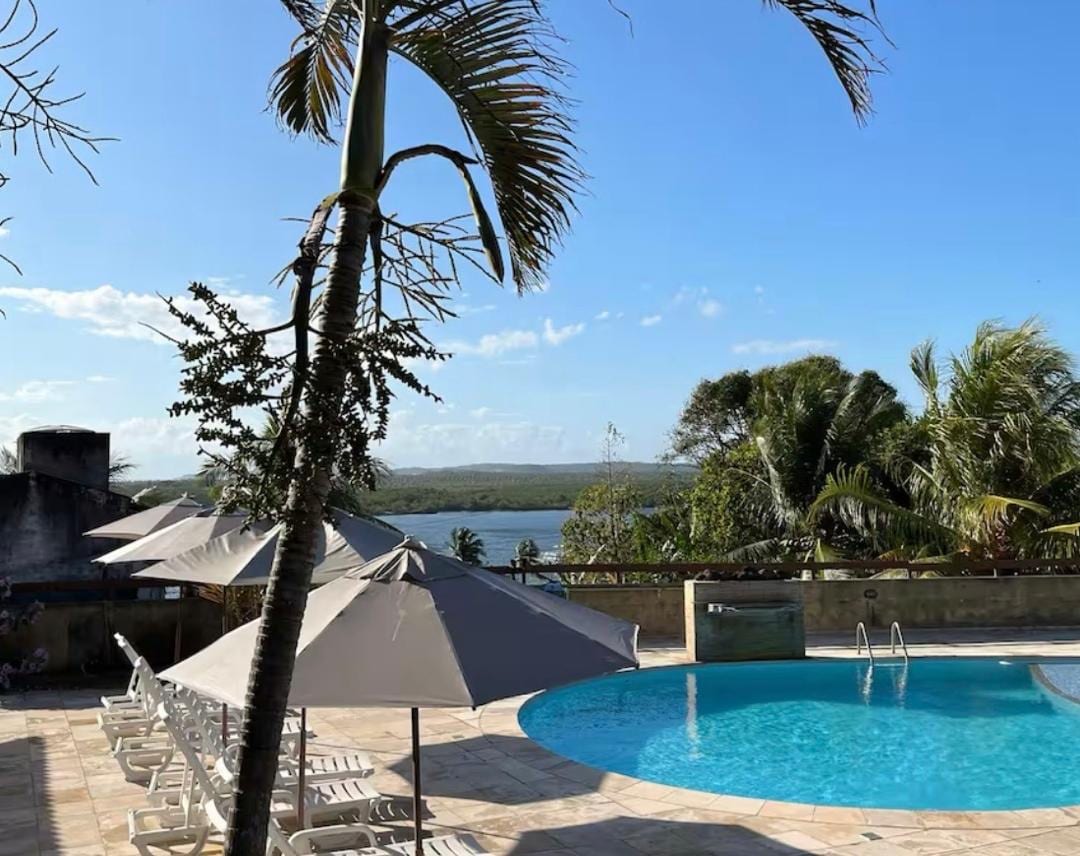 A serene outdoor pool is framed by lush palm trees, with sun loungers arranged beneath large umbrellas. The tranquil water showcases a beautiful blue hue, while a scenic view of the lake and distant hills is visible in the background under a clear sky.