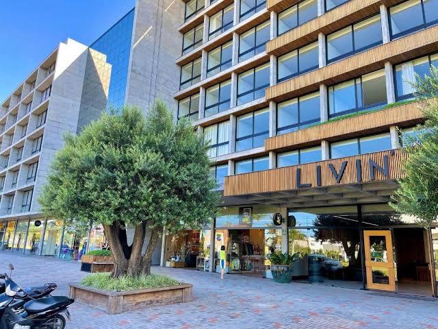 A modern building with a stone and glass façade features large windows along its front. An inviting entryway is framed by a well-maintained tree and planters. The surrounding area includes outdoor seating and spaces for leisurely strolls.