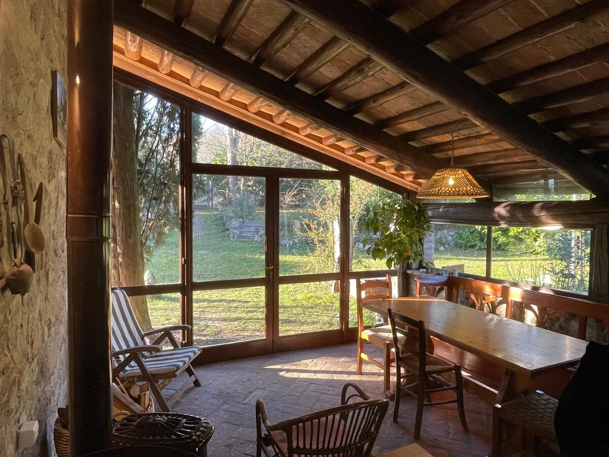 A sunlit room features large glass windows that overlook the green garden. A wooden table is accompanied by several chairs, enhancing the inviting space. Natural light highlights the rustic ceiling and the warm tones of the furniture, creating a serene environment.