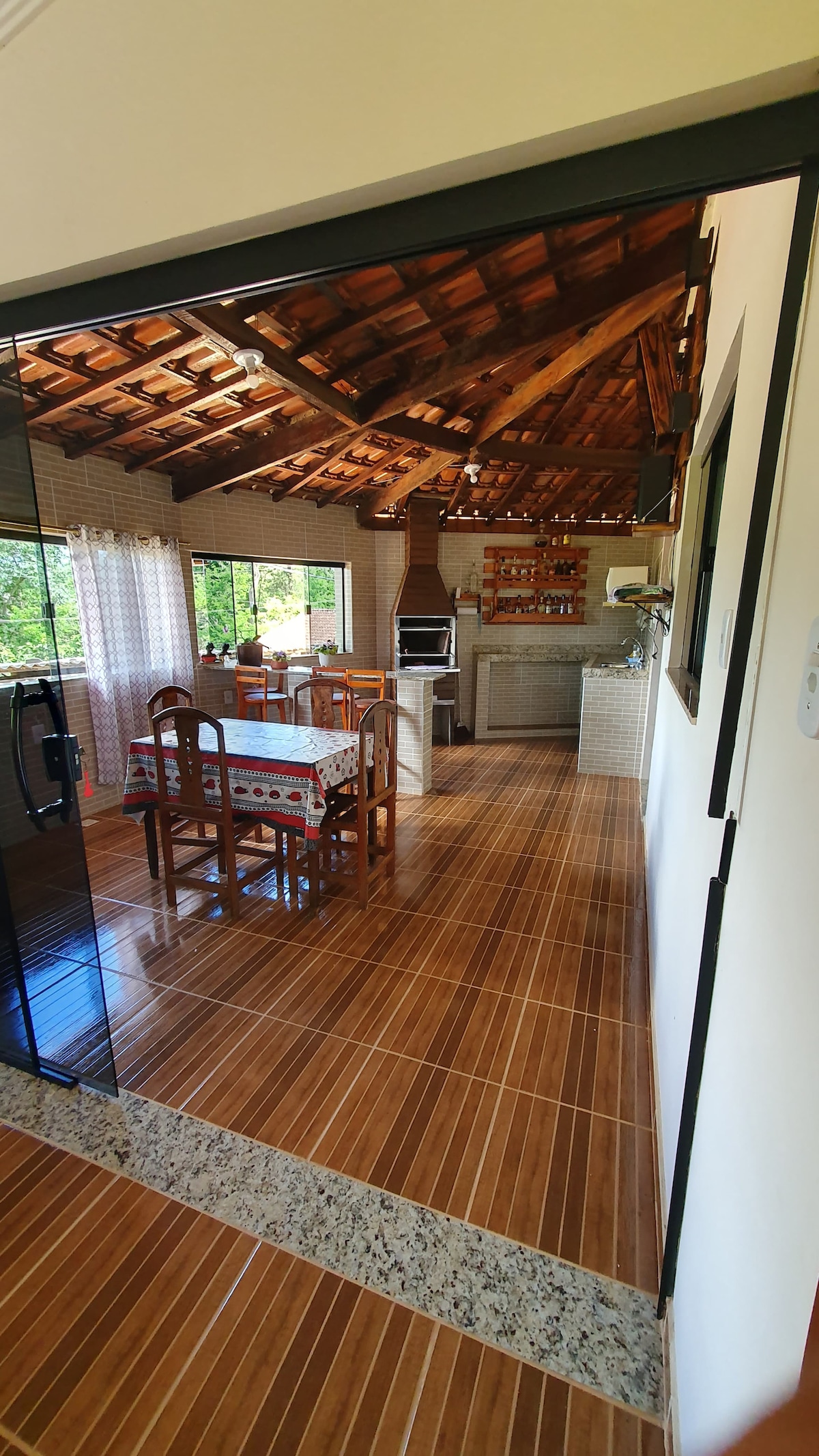 The image captures a spacious dining area featuring a wooden table set for several people, illuminated by natural light from large windows. A rustic kitchen with exposed wooden beams is visible in the background, along with modern appliances and a barbecue area.