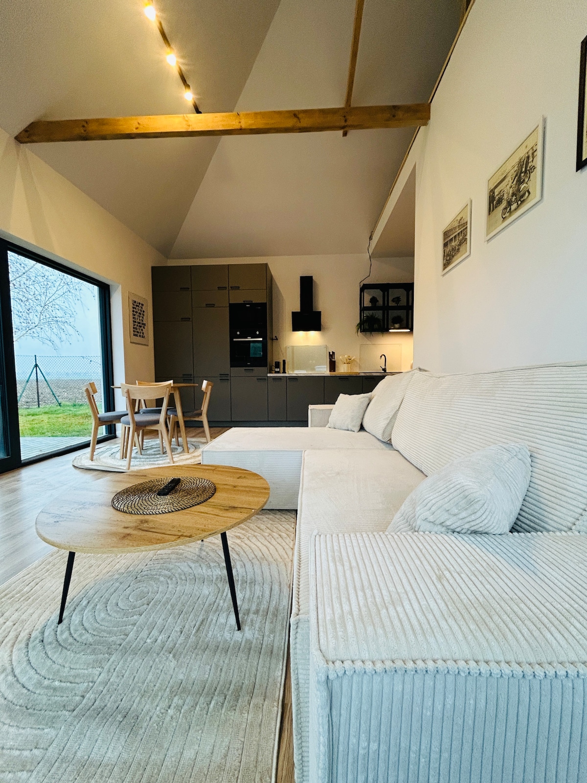 A spacious living area is featured, showcasing a light-colored sectional sofa and a round wooden coffee table. The open kitchen is visible in the background, equipped with modern appliances. Natural light fills the space through large glass doors, offering views of the outdoor garden.