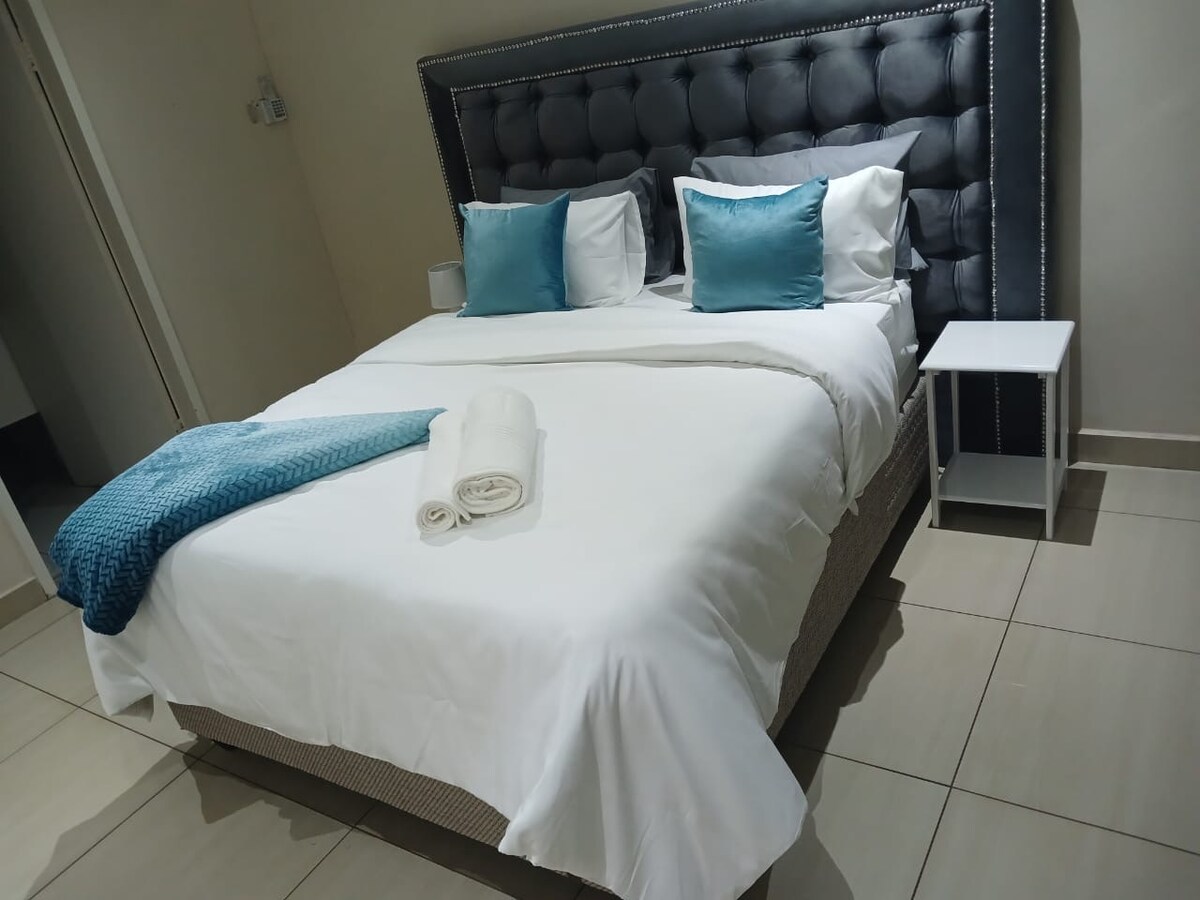 A spacious bed with crisp white linens is adorned with two blue accent pillows. A folded turquoise throw and rolled towels are elegantly placed at the foot of the bed. A modern nightstand sits beside the bed, contributing to a clean and organized appearance.