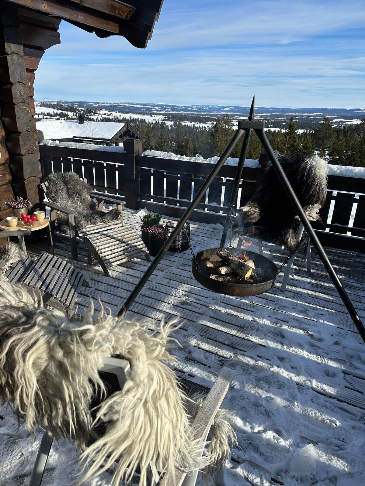 A cozy outdoor space features a fire pit surrounded by chairs and small tables. Soft, fur-like coverings adorn the seating, enhancing comfort. A scenic view of snow-covered hills and trees can be seen in the background under a clear sky.
