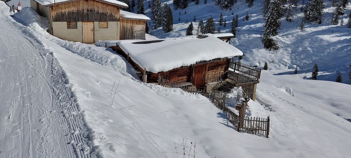 Two rustic wooden cabins are nestled within a snowy landscape, surrounded by evergreen trees. Snow blankets the roofs and the ground, creating a serene winter scene. A wooden fence outlines the path leading down from the upper cabin to the lower one.