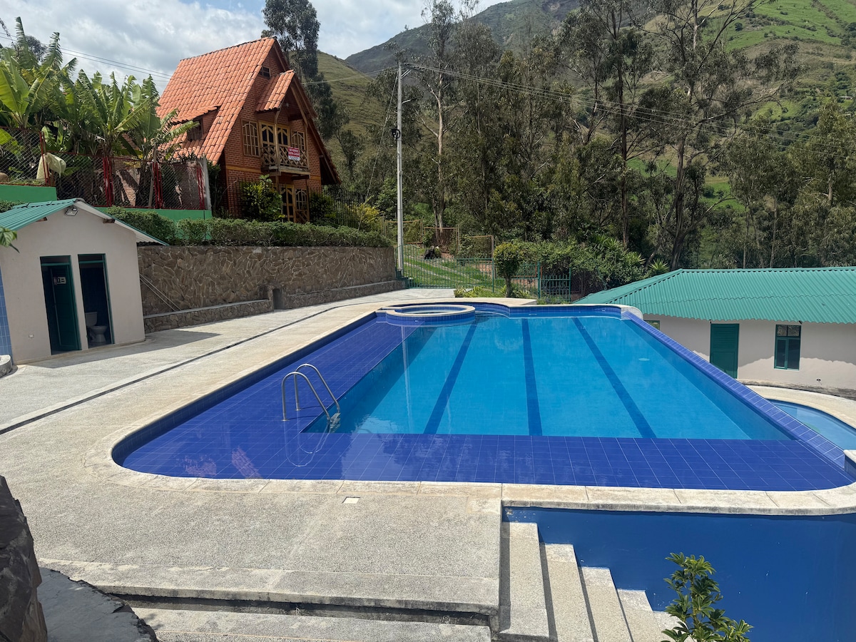 A large swimming pool is set against a scenic backdrop, featuring clear blue water with steps leading into it. Surrounding areas include lounge spaces and greenery, with a rustic lodge visible in the distance under a partly cloudy sky.