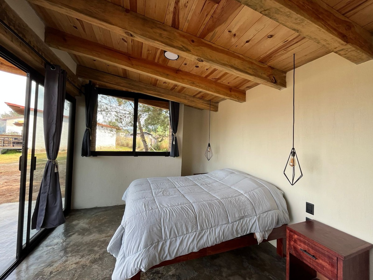 A bright sleeping area features a spacious bed covered with a light quilt. Large windows framed by simple curtains allow natural light to fill the room. The wooden ceiling beams add warmth, while a bedside table sits next to the bed, enhancing the minimalist design.