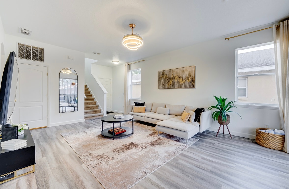 A spacious living area features modern furnishings, including a light-colored sectional sofa and a circular coffee table. A plush area rug softens the floor, while large windows allow natural light to illuminate the room. A decorative plant adds a touch of greenery.
