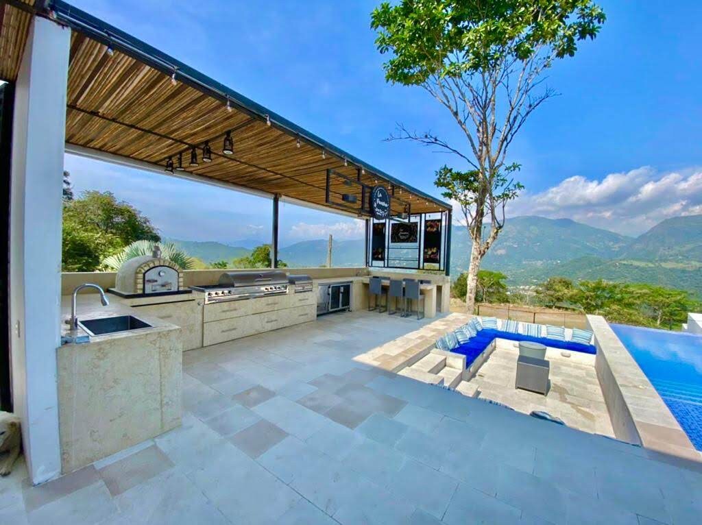 An outdoor kitchen area features a built-in grill and stone countertop, accompanied by bar seating. Nearby, a swimming pool with a jacuzzi is situated against a backdrop of mountains and a clear sky. Lush greenery surrounds the space, providing a natural setting.