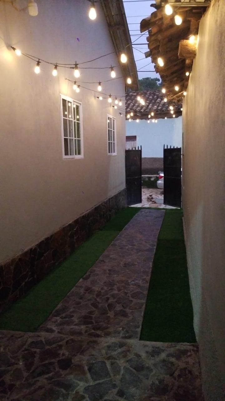 An inviting pathway is bordered by grassy turf and stone tiles, leading towards a black gate. Warm string lights are suspended above, creating a soft glow along the corridor. White walls and small windows frame the space, enhancing the tranquil atmosphere.