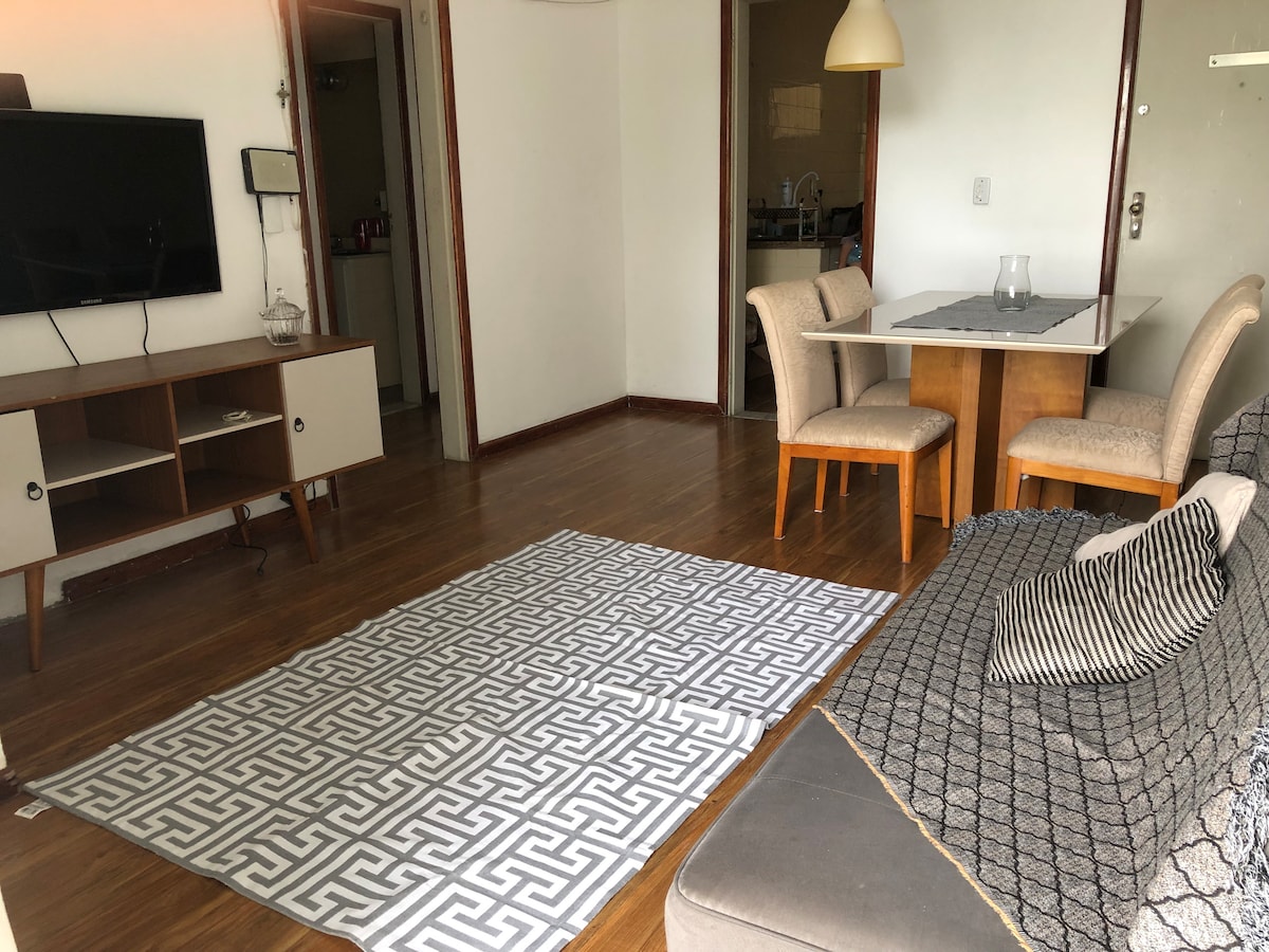 The living area features a comfortable sofa, a patterned area rug, and a wooden dining table with four chairs. A wall-mounted television is visible, along with a soft overhead light. The open layout invites natural light from the nearby entrance.