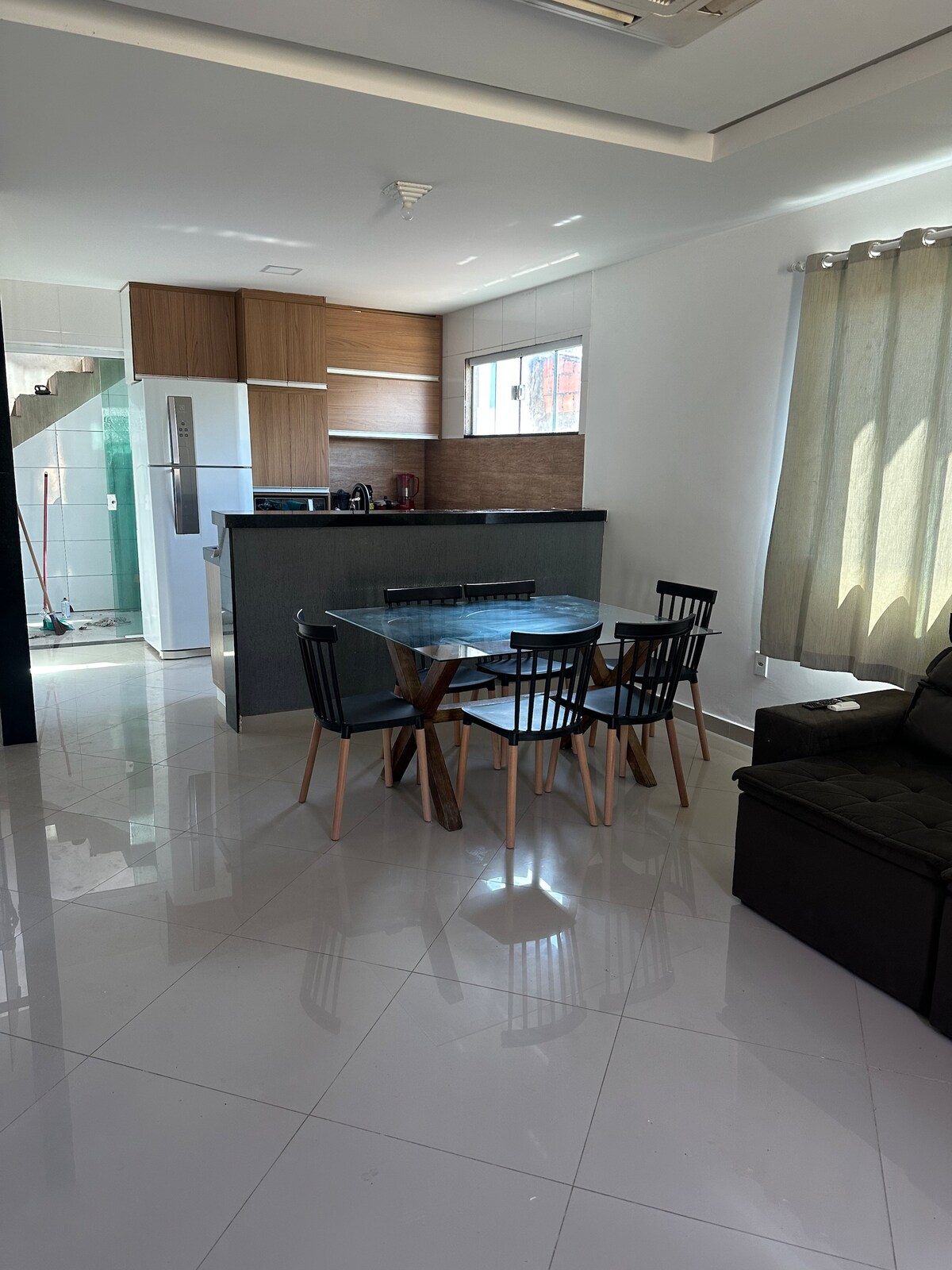 An open-plan dining area features a round glass table surrounded by six black dining chairs. A modern kitchen is visible in the background, showcasing wooden cabinetry and stainless-steel appliances. Natural light enters through large windows, illuminating the glossy tiled floor.