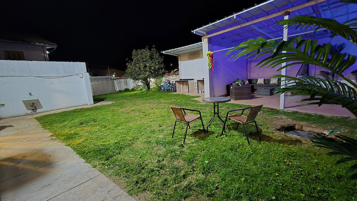 A landscaped outdoor space is illuminated under the night sky. Two chairs are positioned around a small table on the grass, while a covered seating area with modern furniture is seen in the background, adding a relaxed ambiance to the area.