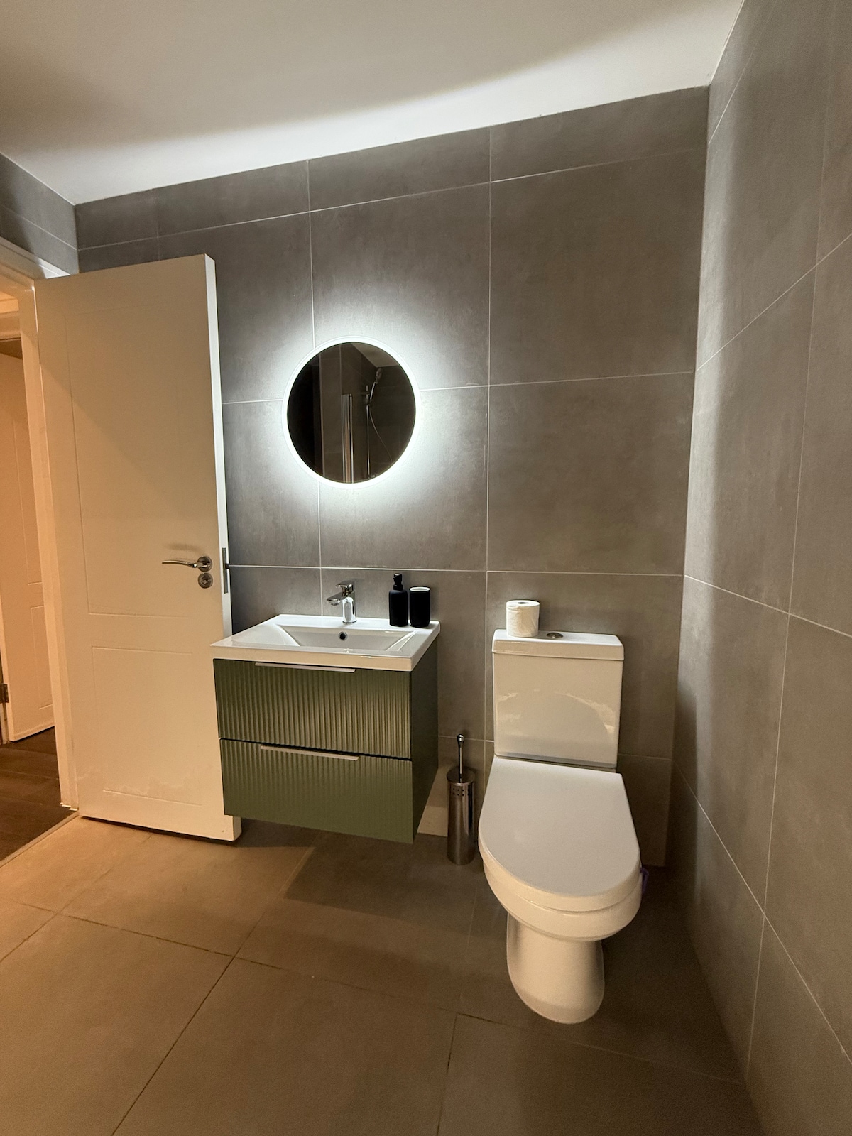 A modern bathroom features a clean, minimalist design with gray tiled walls and flooring. A round mirror with ambient lighting is situated above a compact sink on a green vanity. A toilet is positioned next to the sink, creating a functional space.