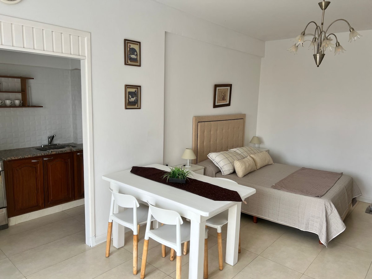 A bright living area is furnished with a spacious bed and a dining table with two white chairs. Soft neutral tones decorate the walls, while framed artwork adds a touch of character. A kitchenette is visible in the background, offering convenient meal preparation options.