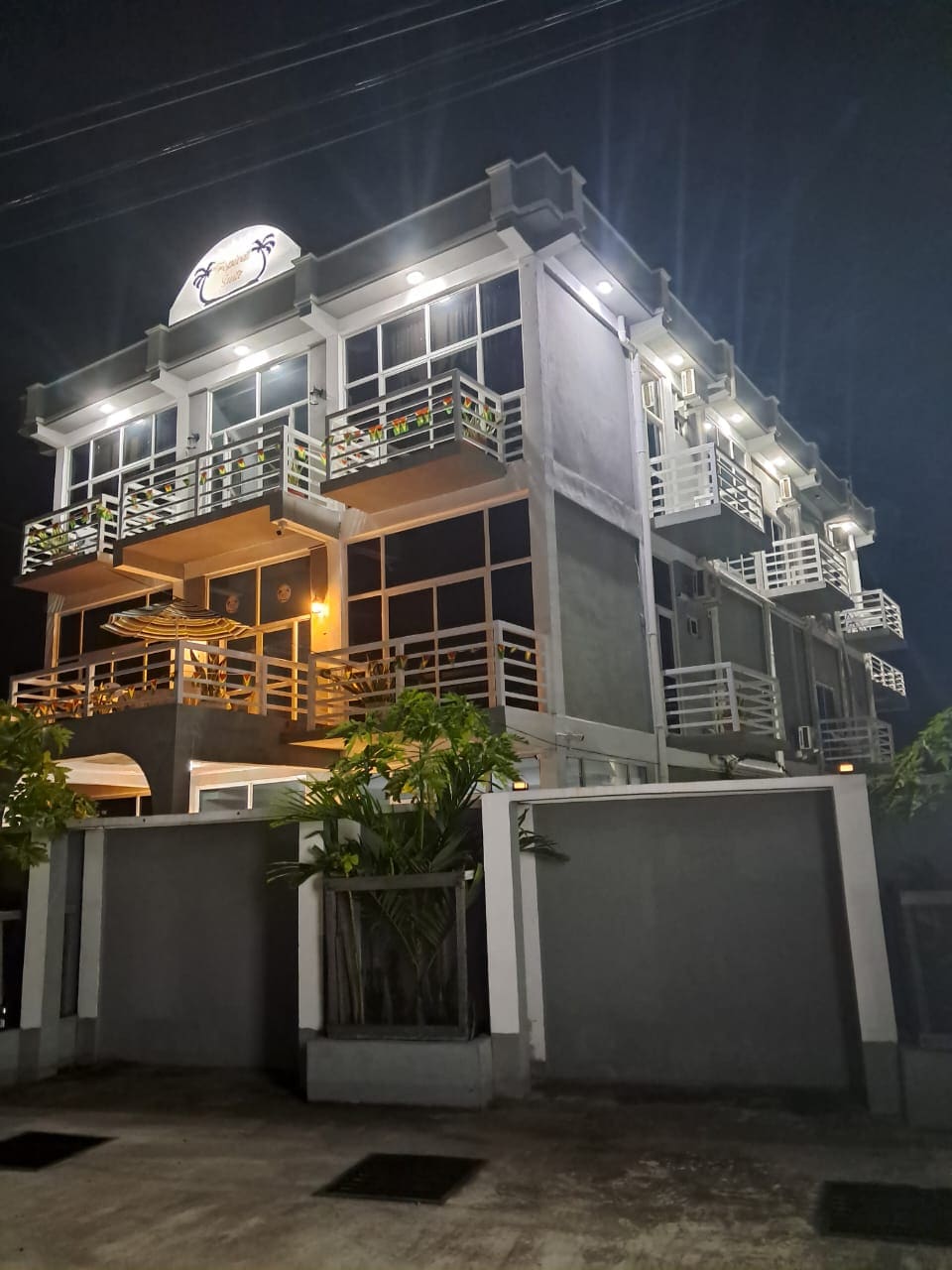 A four-story building is shown at night, featuring multiple balconies with white railings. Warm lighting highlights the entrance area, while surrounding greenery adds a natural touch. Large windows allow a glimpse into the illuminated interior.