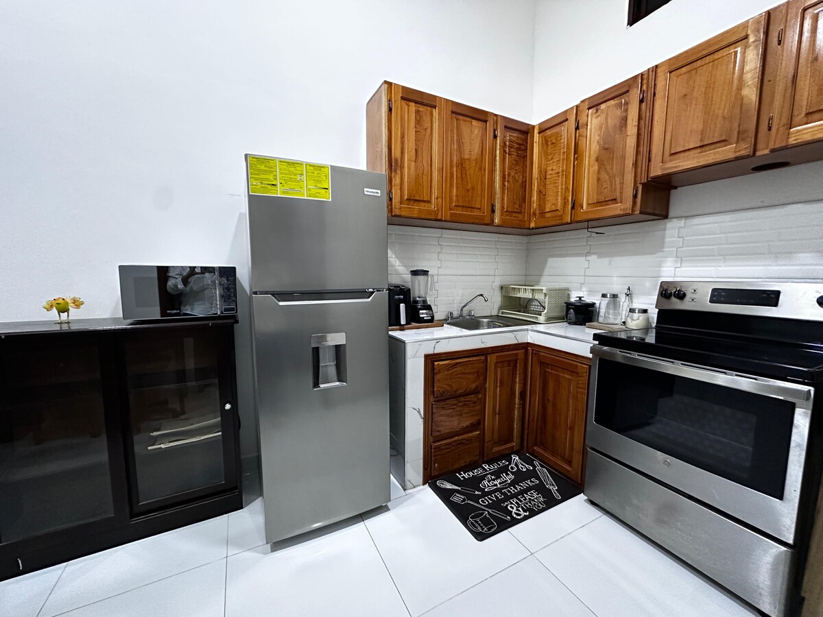 A well-equipped kitchen features wooden cabinetry and stainless steel appliances, including a refrigerator and oven. A blender and microwave are visible on the countertop. The space is complemented by a black mat with a decorative design placed in front of the sink.