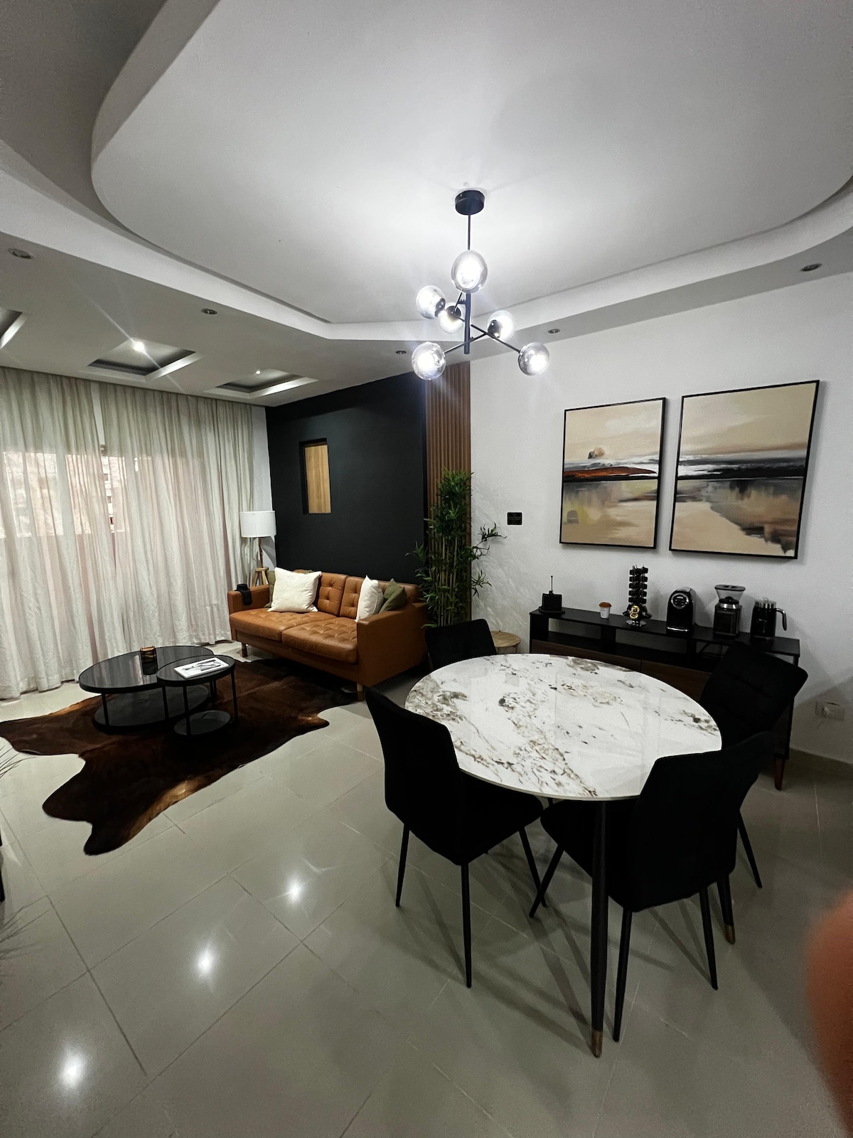 The living space features a cozy sofa paired with a round coffee table on a textured rug. A dining area with a marble table and black chairs is present. Two framed artworks adorn the wall beside bright curtains, creating a balanced and inviting atmosphere.